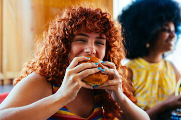 Multiethnic group of friends having meal at 80s vintage diner restaurant, woman biting cheeseburger - Multiracial young people bonding and having fun, eating in an american fast food burger house