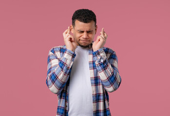 Young man praying with crossed fingers on pink background. Guy begs someone
