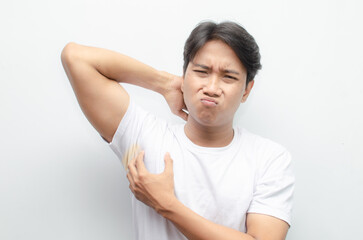 asian man feels bad after smelling his armpit with yellow stain on his white tshirt. young asian man smells his bad odor armpit with disgusted face.