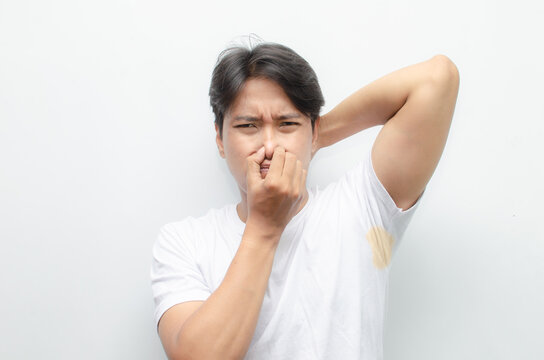 asian man feels bad after smelling his armpit with yellow stain on his white tshirt. young asian man smells his bad odor armpit with disgusted face.