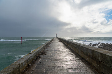 Fototapeta premium Le phare du Raoulic se reflète partiellement sur le sol mouillé du môle, où les reflets des nuages et du soleil dansent en Bretagne.