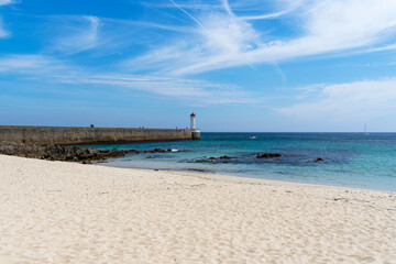 Vue sur le môle et le phare du Raoulic, avec en premier plan une plage de sable et des rochers, le tout sous un ciel bleu légèrement voilé en Bretagne.