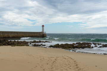 Au bout du môle se dresse le phare du Raoulic, tandis qu'au premier plan se trouvent plage de sable et rochers, le tout sous un ciel partiellement couvert en Bretagne.