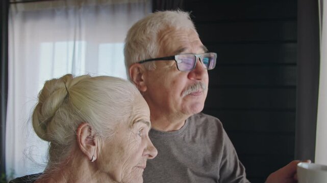 Close-up of senior Caucasian mand and woman standing together by window at home, drinking cups of tea, hugging, gossiping, chatting, laughing while enjoying happy times in retirement