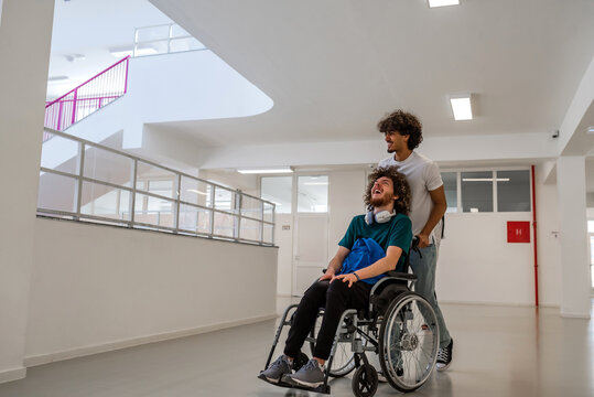 A happy college students in university hallway, a classmate pushing a friend in a wheelchair as they walk down the school corridor or male patient smiling with friendly caregiver in hospital corridor.