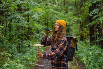 Young woman holding a large mushroom in one hand while using her smartphone to identify it, standing on a forest trail surrounded by lush greenery.  © wifesun