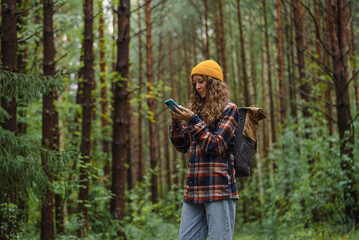 Fototapeta premium Young woman in a plaid shirt and beanie checking her smartphone while hiking through a forest, blending technology with outdoor adventure. 