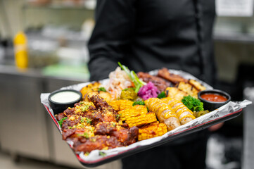 A waiter holding a tray with assorted grilled foods, including corn, sausages, and ribs, in a restaurant setting. The background features a cozy dining area with warm lighting and wooden furniture