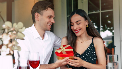 Happy Valentine's day, anniversary, and birthday. A man presents a beautifully wrapped gift in shape of heart to a smiling woman as they enjoy pastries and wine together in restaurant.