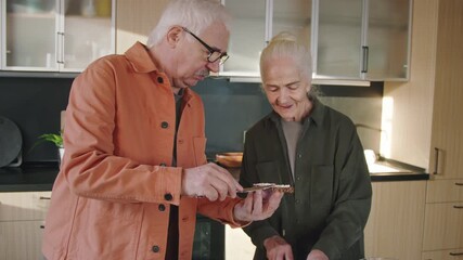 Medium tilt-down shot of senior Caucasian man buttering piece of bread and debating healthy food choices with wife, who is cutting vegetables for salad in kitchen of spacious family home
