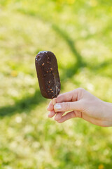 Close-up of chocolate covered ice cream on stick in woman hand against background of grass in summer park. Summer cooling treat. Summer break time