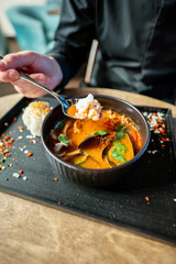 A person holding a spoon over a bowl of vibrant soup with vegetables, served on a dark tray with a side of rice. The scene is set on a rustic wooden table, creating a cozy and inviting atmosphere.