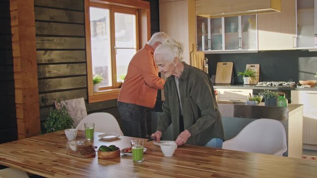 Full shot of senior Caucasian couple laying table for healthy breakfast in morning, bringing sandwiches, salad, smoothies and muffins, while joyfully chatting, joking and smiling