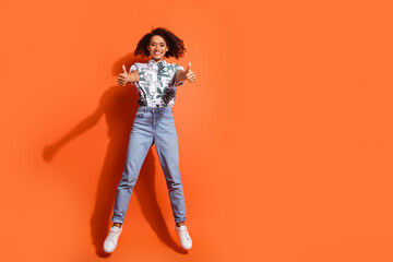 Full body photo of american woman in jeans and summer stylish shirt jumping trampoline positive rating thumbs up isolated on orange color background