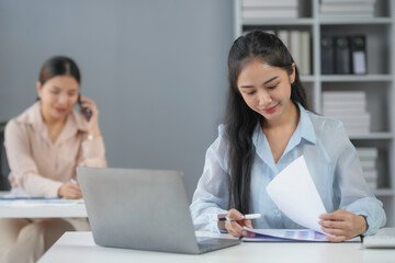 Obraz premium Focused asian businesswoman is working with financial documents at her desk while her colleague is making a phone call
