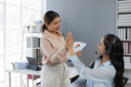 Two happy businesswomen giving high five while holding financial report with chart, celebrating successful project and teamwork in modern office