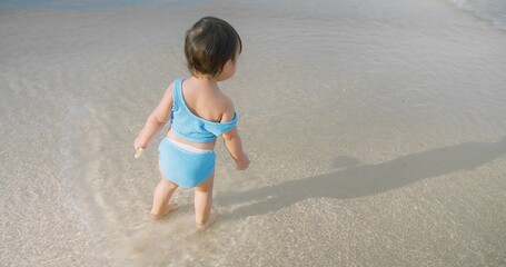 A young Toddler child wearing a blue swimsuit stands in shallow beach water, casting a shadow on a sunny day summertime