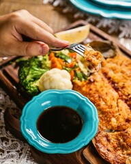 Man eating breaded fish fillet using fork with vegetables and sauce on wooden board