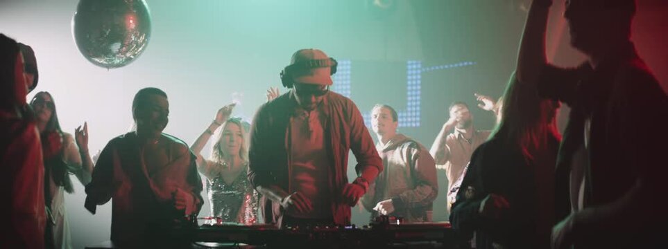 Slow-motion shot of a DJ playing music in club, attractive women dancing around. Nightlife, modern music and entertainment concept