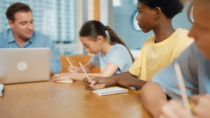 Young smart teenager writing and talking about topic at classroom. Diverse highschool student taking a note in other notebook while sitting with multicultural friends. Creative education. Edification.