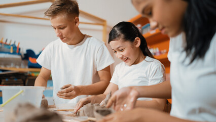 Handsome student dipping hand in to water to soften clay at art classroom. Group of diverse children working or modeling vase. Happy boy put water to young girl while laugh with happy. Edification.