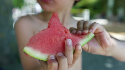 Boy eagerly eating a slice of watermelon, eyes closed in enjoyment, shirtless and outdoors,...