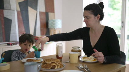 Mother and son having breakfast together at a wooden table, mother pouring syrup onto the boy's pancakes, creating a warm and family-oriented atmosphere