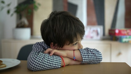 Thoughtful boy in checkered shirt resting his head on folded arms on a wooden table feeling pensive, with a background featuring blurred indoor elements and a touch of natural light