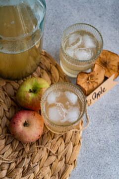 Jug and two glasses of fresh apple juice on a table mat with fresh apples, dried apple slices and a tag with the word apple
