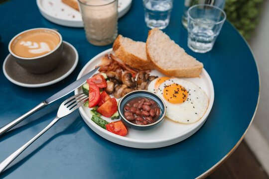 Traditional English breakfast with baked beans,  bacon, fried eggs, mushrooms tomatoes, cucumber and bread with a cappuccino and glass of water