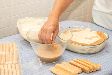 A woman prepares tiramisu dessert in a glass bowl with a mixing bowl on the side in a home kitchen. Step by step recipe. High quality photo