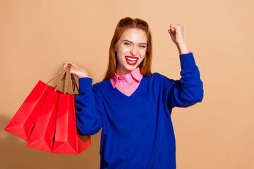 Portrait of pretty young lady hold bags raise fist wear blue sweater isolated on beige color...