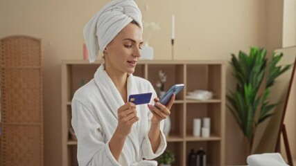 A young attractive blonde woman in a spa wellness room checks her phone while holding a credit card, wrapped in a white towel, surrounded by elegant indoor decor.