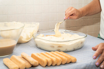 A hand spreads creamy mascarpone over soaked ladyfingers in a glass dish, finalizing a step in making tiramisu, an Italian dessert.