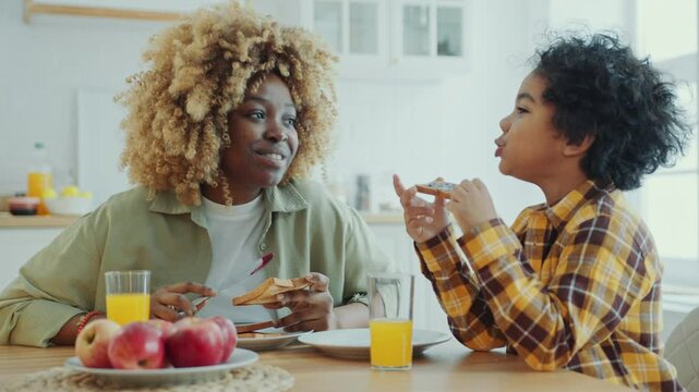 Happy African American mother making jam toast, smiling and talking with cute little son eating breakfast in the kitchen at home