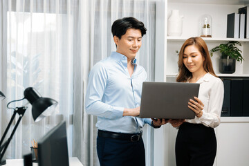 Smiling Asian coworkers standing while talking planning marketing report project on laptop casual day at modern office. Analyzing successful customer need in new social media online channel. Infobahn.