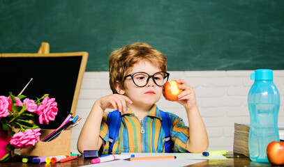 Healthy school breakfast or lunch for kids. Cute child boy eating apple during lunch break in classroom. Little schoolboy in glasses at desk in front of chalkboard eating apple. Tasty food for pupils.