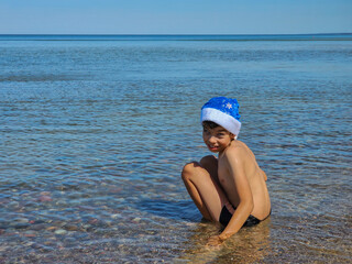 boy sitting in water in santa hat, childhood on trip, child in water, swimming in sea on new year, christmas, child on christmas at sea