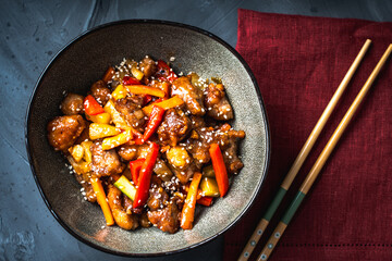 Pork in sweet and sour sauce in a bowl on a dark background with Chinese chopsticks and a burgundy napkin on a dark gray background.