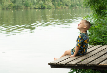 A small handsome boy, with emotional facial expressions, 4 years old, sits on a wooden bridge by the river, against the backdrop of green plants and breathes fresh air. Relax. Place for text.