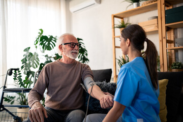 Obraz premium A cheerful nurse in blue scrubs takes the blood pressure of an elderly male patient during a home visit, capturing a moment of attentive health care service.