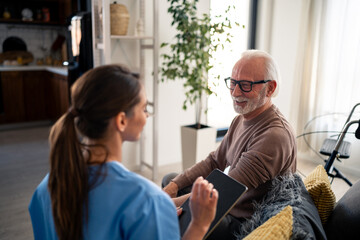 A joyful female nurse in blue scrubs engages with an elderly male patient in his home, discussing...