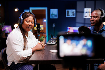 Talk show hosts in neon-lit home studio using professional equipment to create high quality content. Camera positioned on tripod films every move of african american couple doing a podcast episode.