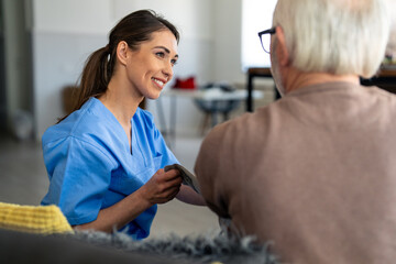 Fototapeta premium Smiling female nurse in blue scrubs measures the blood pressure of an older male patient within the comfort of his home environment.