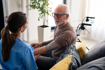 Obraz premium A smiling female nurse in blue scrubs attentively measures the blood pressure of a joyful elderly man seated in a cozy apartment setting.