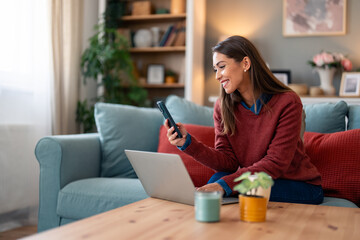 A Caucasian woman in casual attire sits on a couch, engaged in using a laptop while simultaneously...