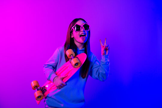 Photo of charming positive small girl wear hoodie holding skateboard showing v-sign isolated neon violet color background