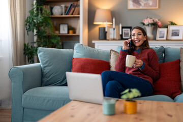 Obraz premium A cheery Caucasian woman in casual attire balances work and communication, expertly handling a laptop and smartphone while seated comfortably on her living room sofa.
