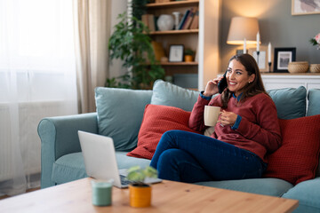 Obraz premium A cheerful Caucasian woman relaxes on her couch, adeptly juggling a conversation on smartphone while navigating a laptop, her casual attire suggesting comfort during her productive afternoon indoors.
