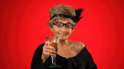 Smiling portrait of fun elderly old woman drinking wine and smiling wearing wig and glasses isolated on red background.
